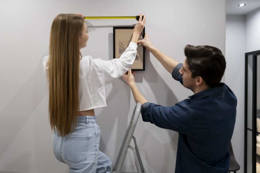 Smiling couple hanging a picture frame on a freshly finished drywall surface in a modern living room.