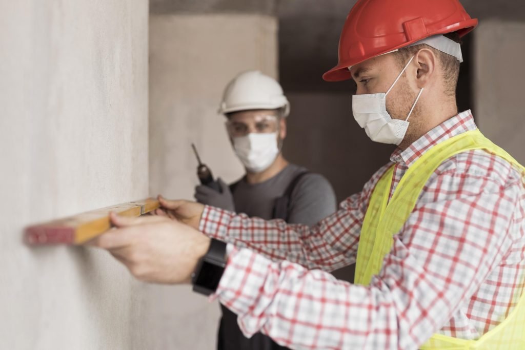 Two workers wearing protective masks while handling drywall installation in an indoor construction area.